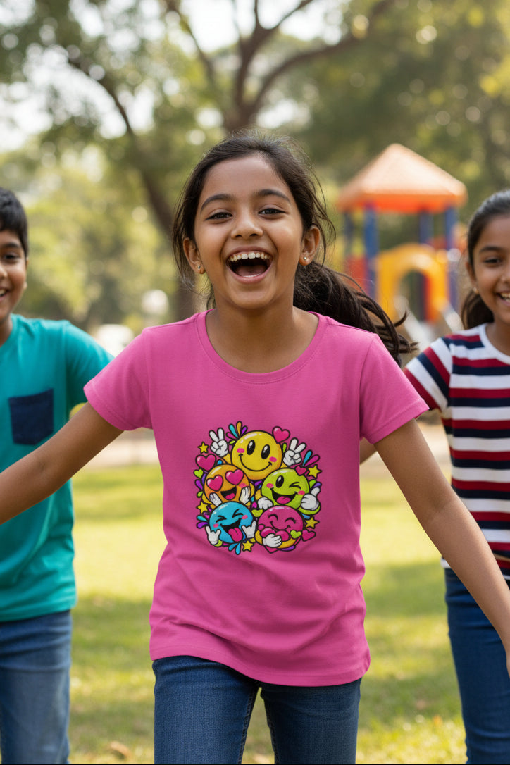 A pink short-sleeve crewneck t-shirt featuring a colorful graphic print of cartoon emoji faces surrounded by hearts and flowers on the chest.